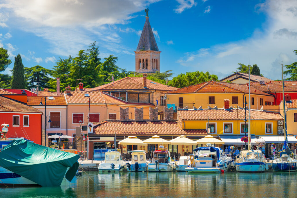 Boats in Fažana harbour below colourful waterfront houses and a church bell tower.