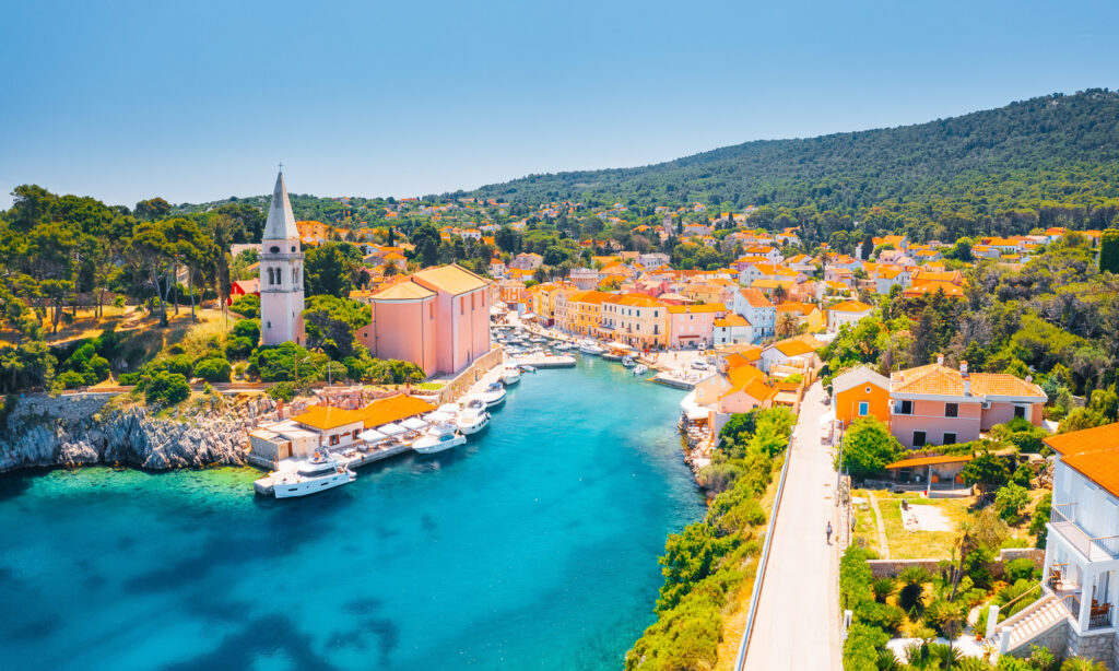 Aerial view of Veli Lošinj harbour, with boats, pastel waterfront buildings and wooded hills.