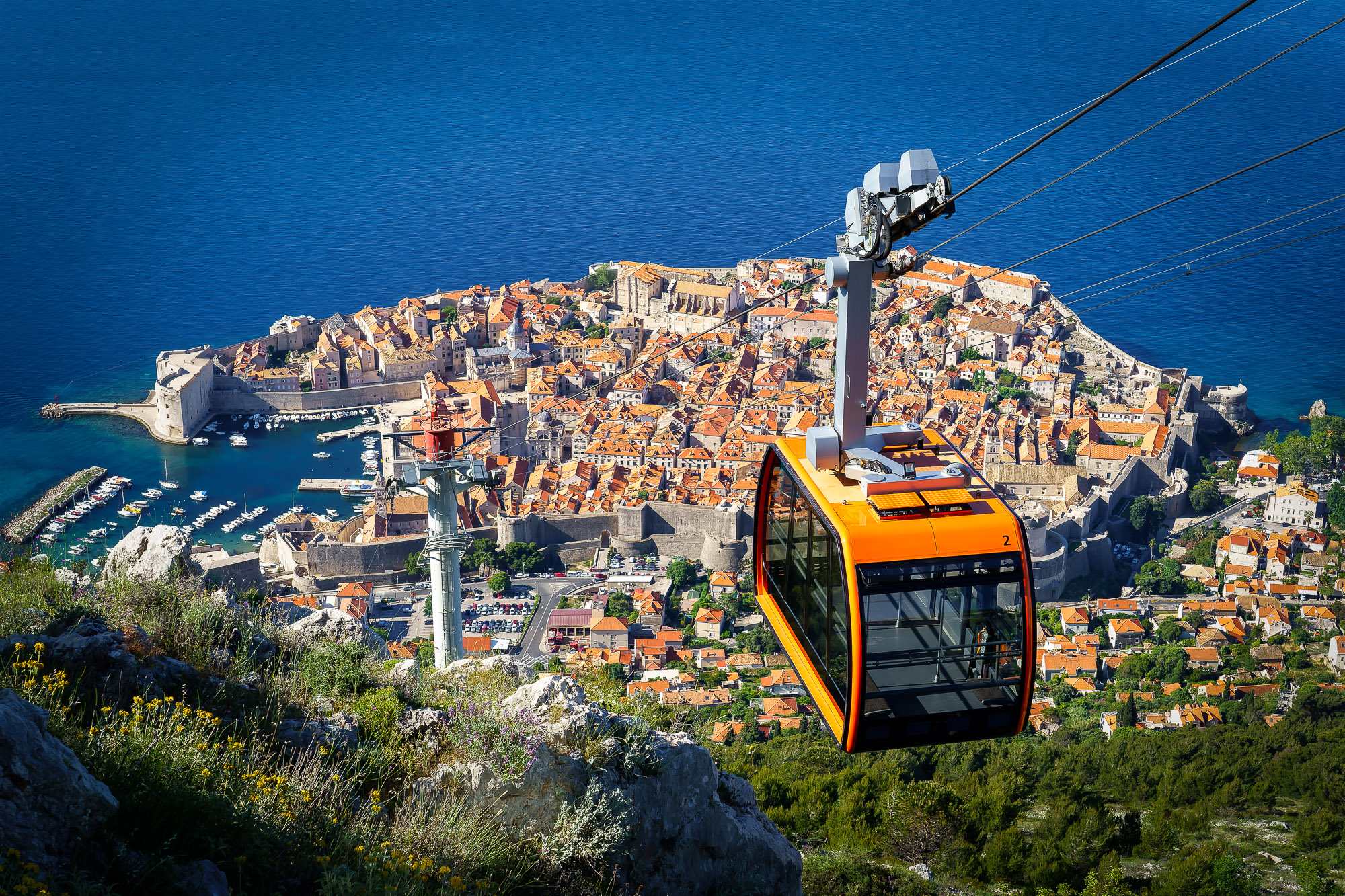 An orange cable car passes above Dubrovnik Old Town, its terracotta roofs bordered by the Adriatic Sea.