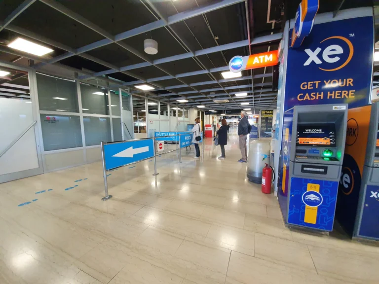 Airport terminal with shuttle-rental directions, taxi departures signage and a currency exchange ATM