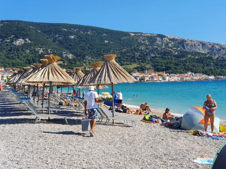 Pebble beach with thatched umbrellas and swimmers, backed by Baška and a steep mountain ridge.