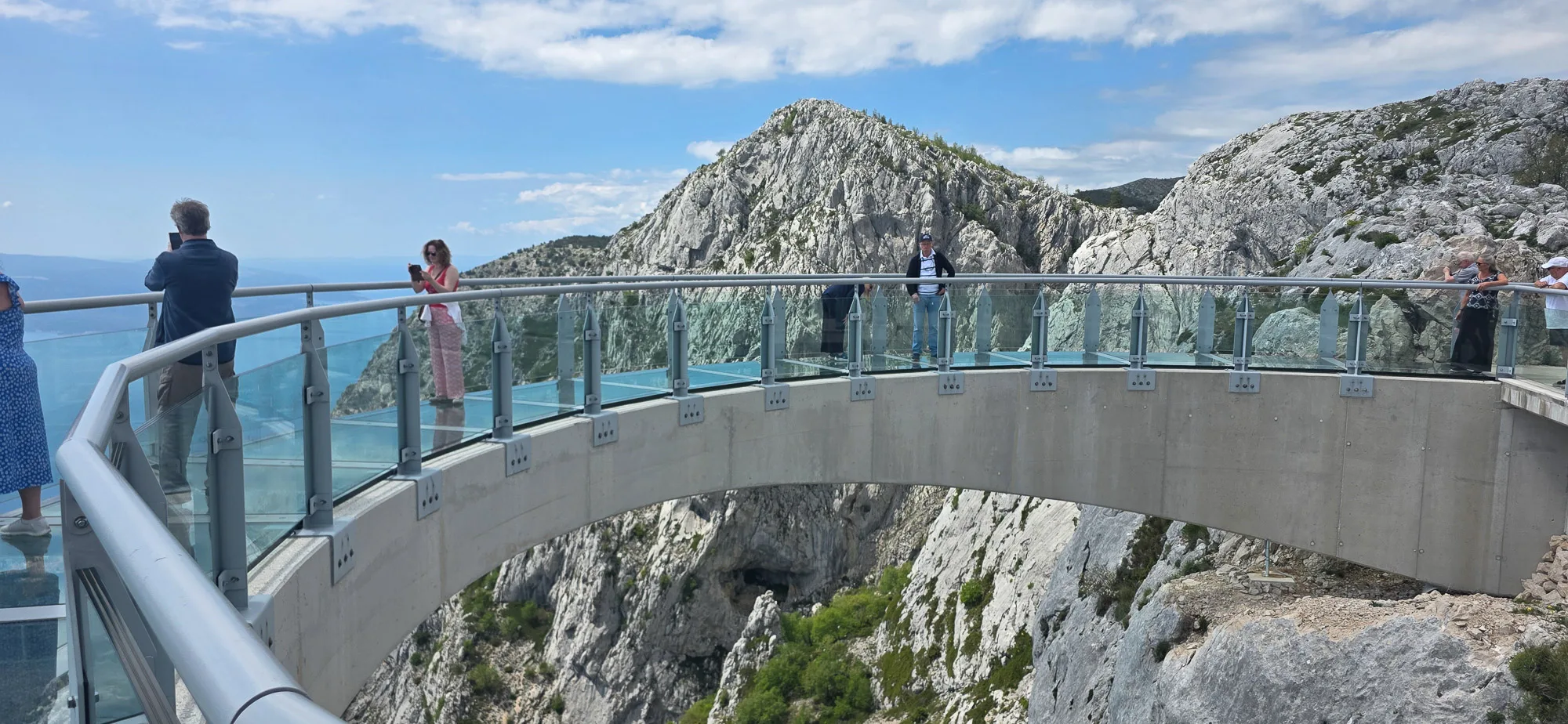 Visitors stand on the curved glass Skywalk Biokovo above rugged mountain cliffs, with the Adriatic visible beyond.