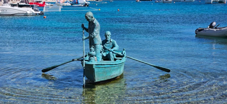 Two bronze figures sit in a small rowing boat in clear harbour water, with sailboats in the background.