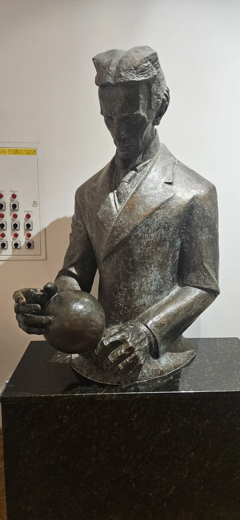Bronze statue of a man in a cap holding a sphere against a plain indoor wall