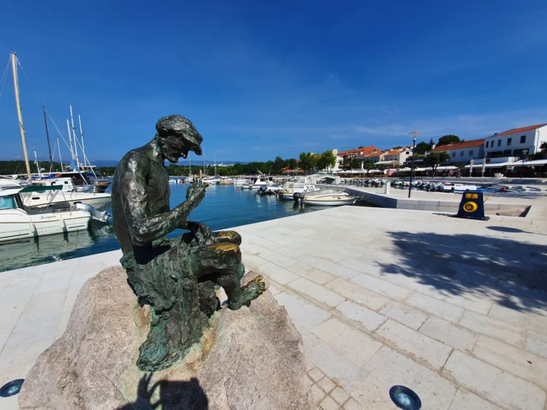 A seated bronze statue overlooks a marina filled with boats beside a sunlit waterfront promenade.