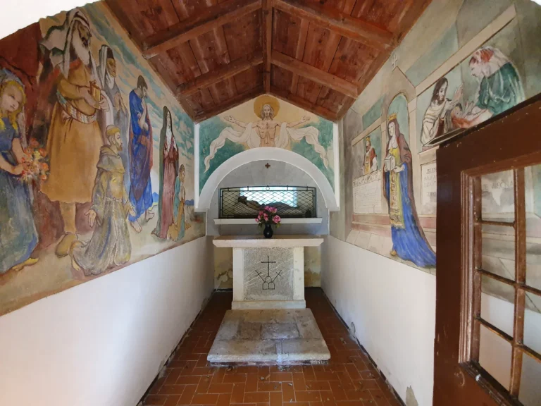Small chapel interior with painted religious frescoes, a stone altar and a wooden ceiling