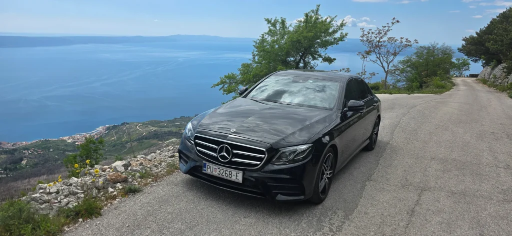 Black car parked on a coastal road above the sea, with a wide view of the shoreline and distant hills.