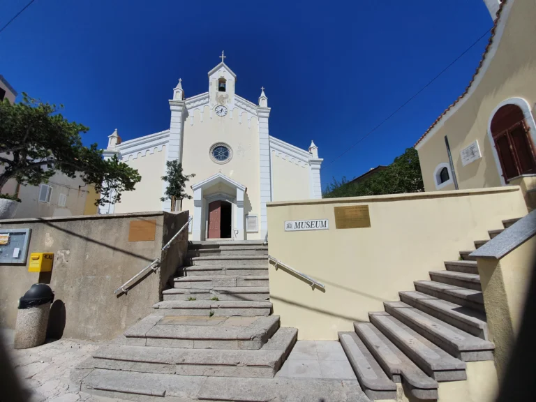 White church with a clock and bell tower above stone steps, beside a wall marked “Museum.”