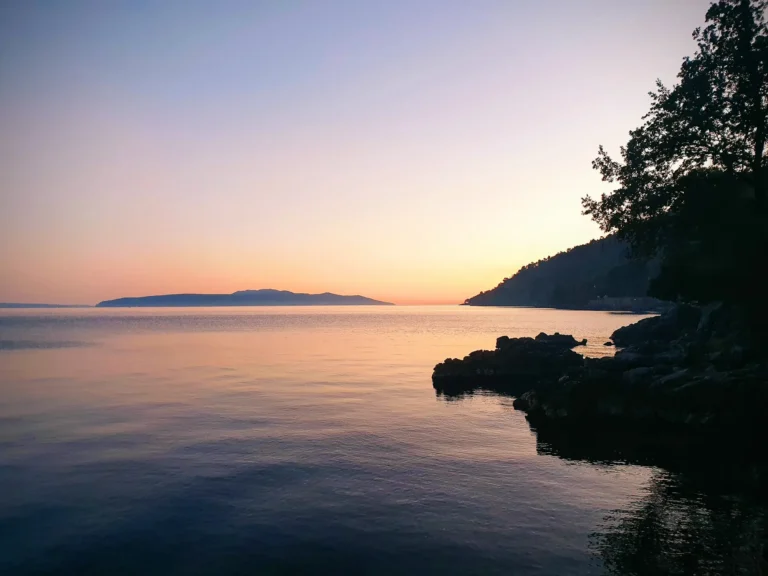 Sunset over calm water on the Croatian coast, framed by a dark rocky shore and trees.