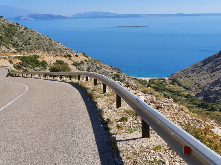 A winding coastal road descends past rocky hills toward a small beach and the blue Adriatic Sea in Croatia.