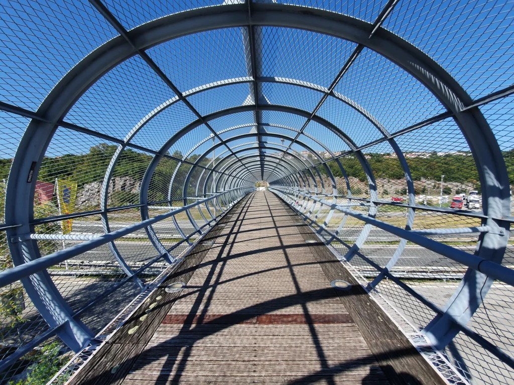 A wooden walkway crosses beneath the curved metal mesh roof of an enclosed pedestrian bridge.