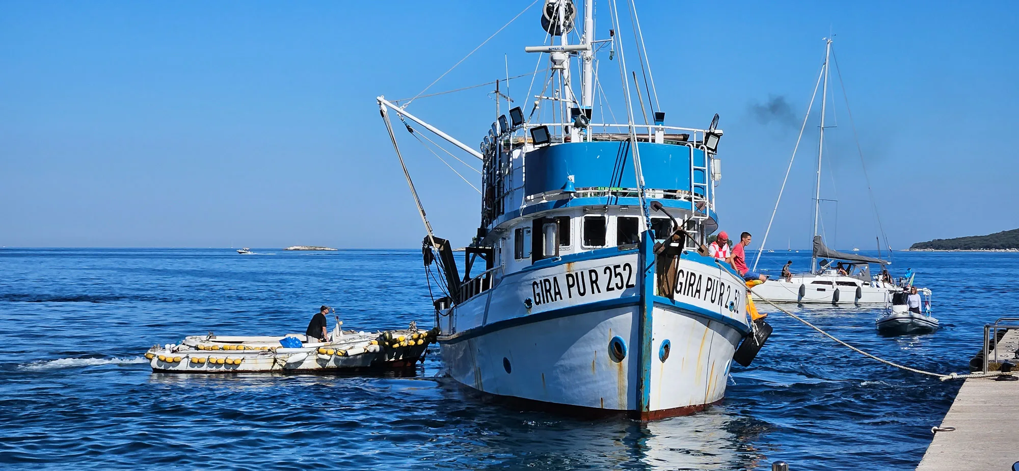 A large fishing boat moored beside smaller boats at a Croatian harbour on a clear day.