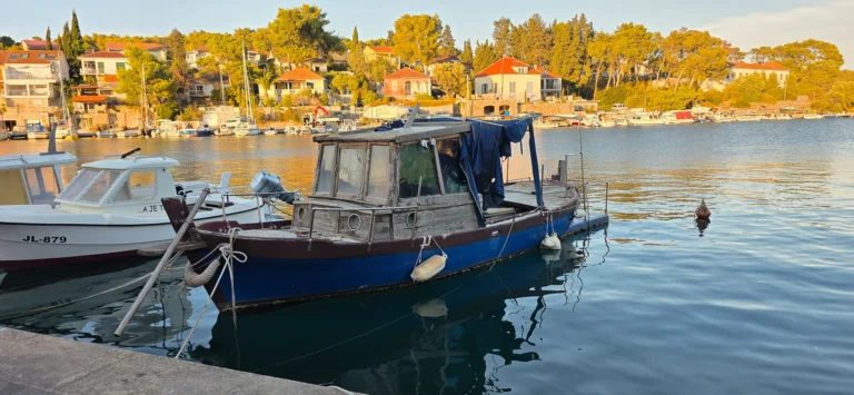 A blue wooden fishing boat and smaller motorboats moored beside a tree-lined Croatian waterfront.