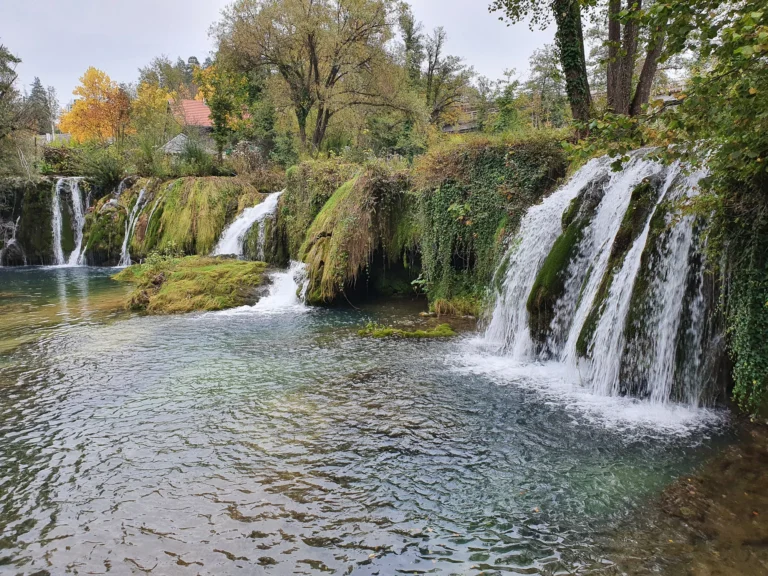 Several waterfalls cascade over moss-covered rocks into a clear river beneath autumn trees.
