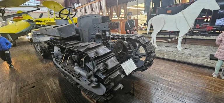 A tracked military vehicle on display in a museum, with an aircraft and historic train exhibits behind it
