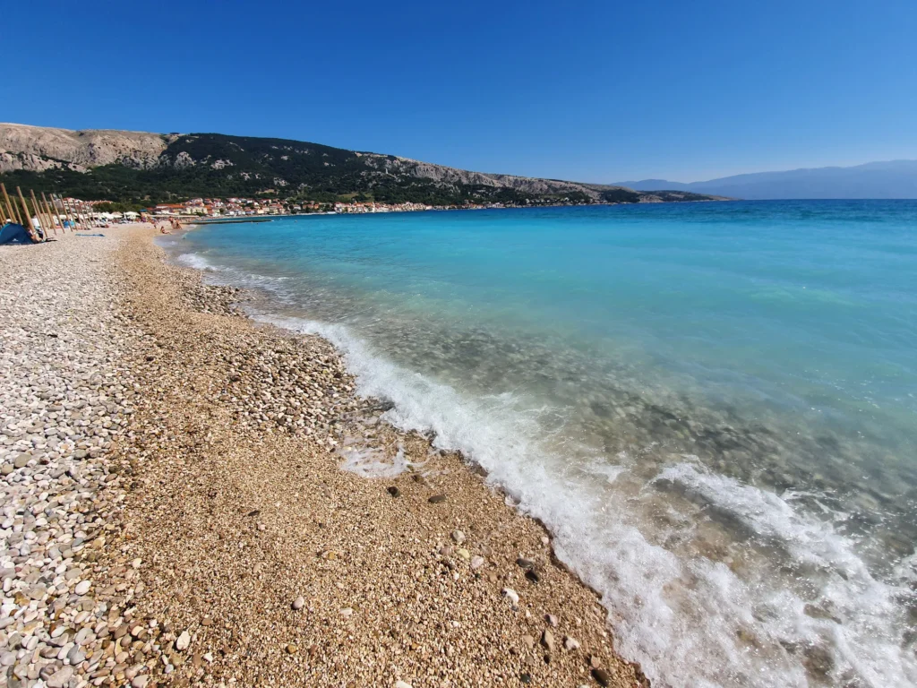 A broad pebble beach meets clear turquoise water, with a coastal town and rocky mountains in the distance.
