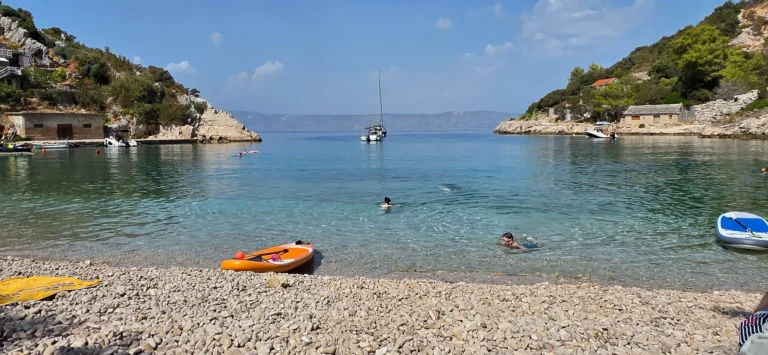 Pebble cove on Croatia’s coast with swimmers, a paddleboard and a sailboat offshore.