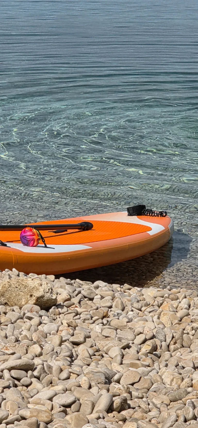 An orange paddleboard rests on a pebble shore beside clear, shallow water in Croatia.