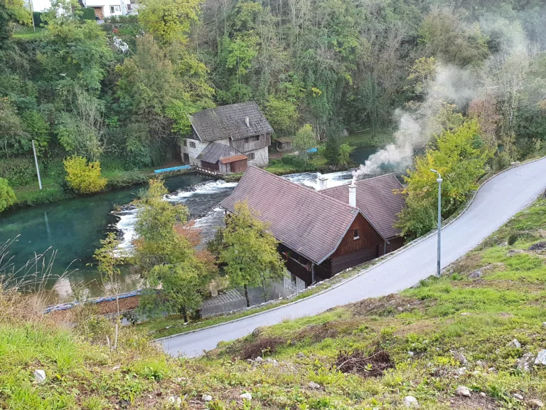 Old mill buildings beside a fast-flowing river and small rapids, viewed from a hillside road.