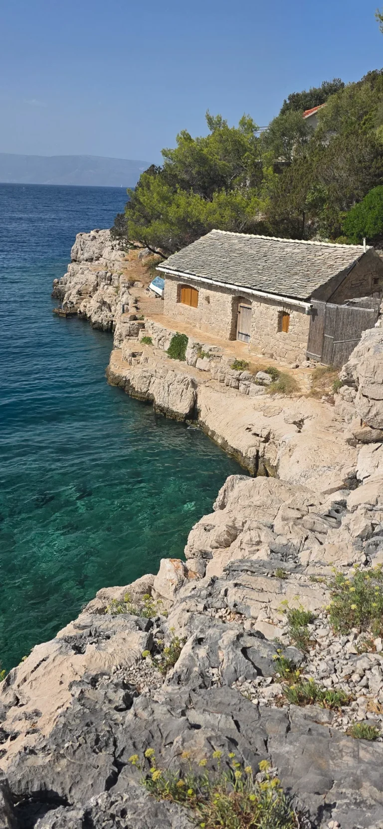 A stone cottage stands on a rocky Croatian coast above clear turquoise water, with pine trees behind it.