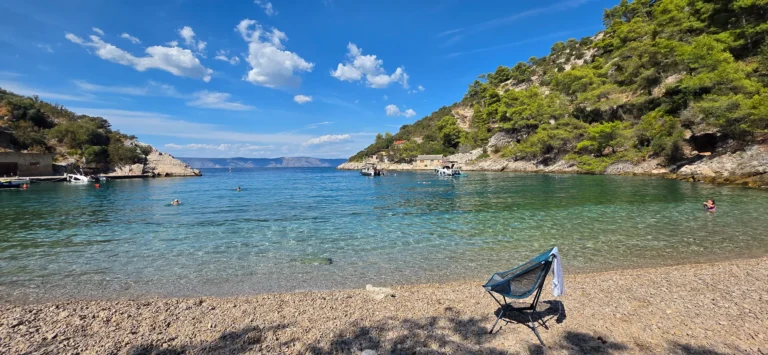 A rocky pebble cove in Croatia with clear turquoise water, small boats and wooded hills.