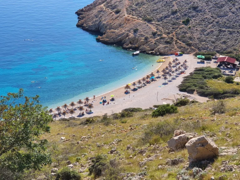 A pebble beach with thatched parasols sits below rocky slopes beside clear blue water in Croatia.