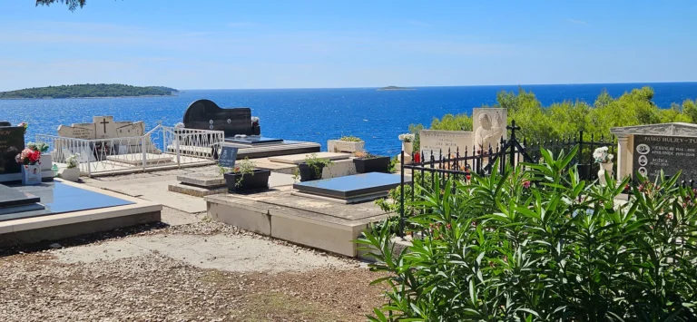 Rows of gravestones in a coastal cemetery overlook the blue sea and small islands.