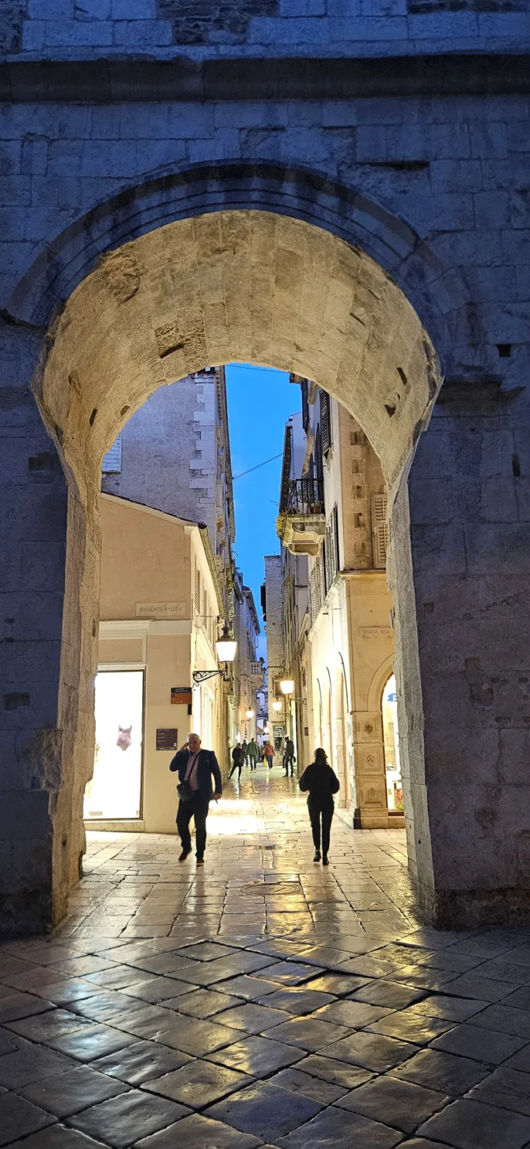A large stone arch frames a narrow pedestrian street with historic buildings and evening lights.