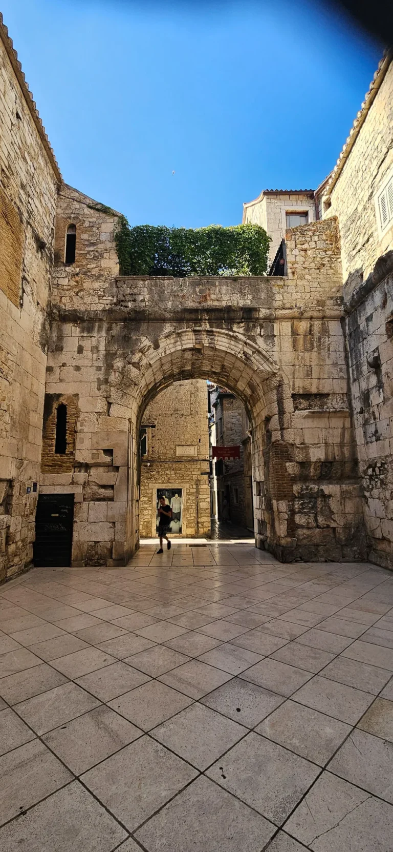 A large stone arch frames a narrow passage between old stone buildings in a paved courtyard.