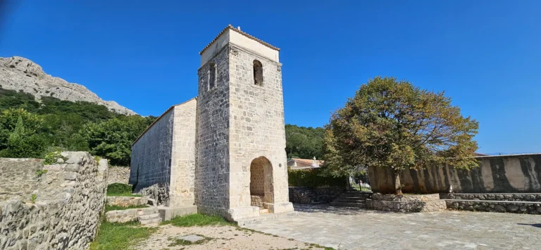 Stone church with a square tower in a walled courtyard beneath a rocky, forested mountain