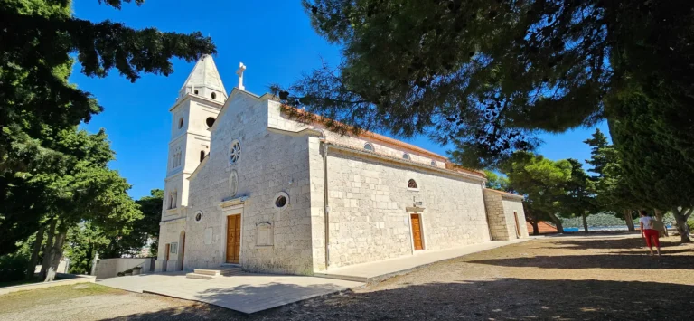 A pale stone church with a bell tower stands among pine trees near the coast in Croatia.