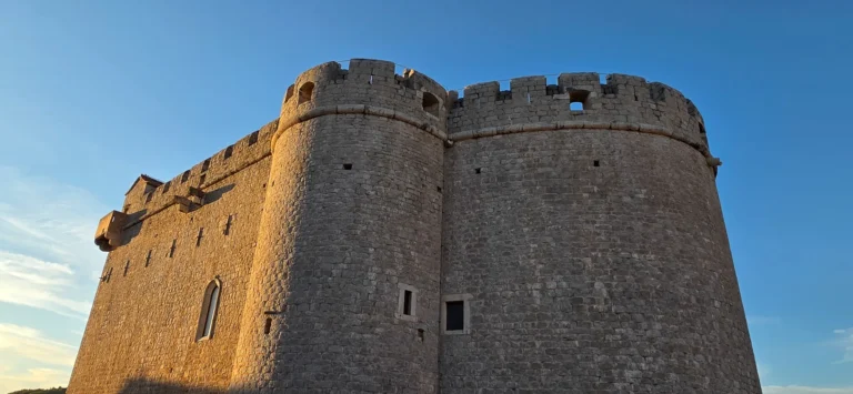 A massive stone fortress with rounded towers catches warm light beneath a clear blue sky in Croatia.