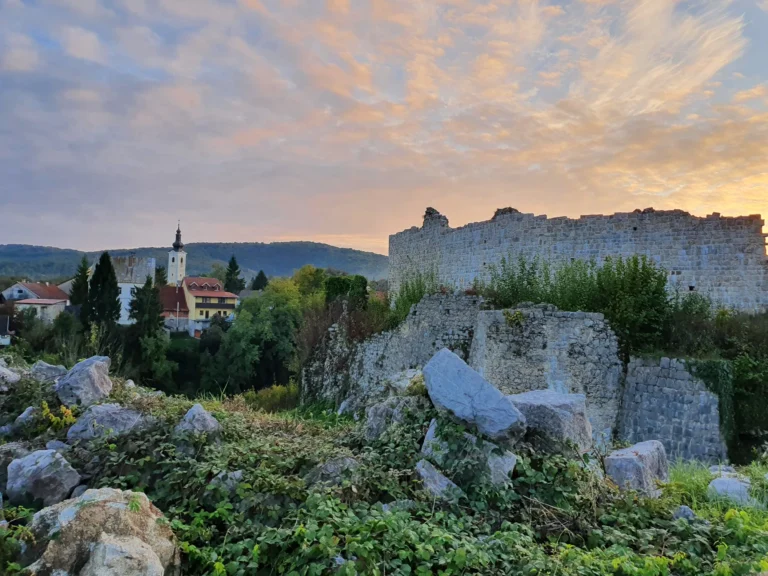 A weathered stone fortress wall overlooks houses and a church tower beneath a pastel sunset sky.