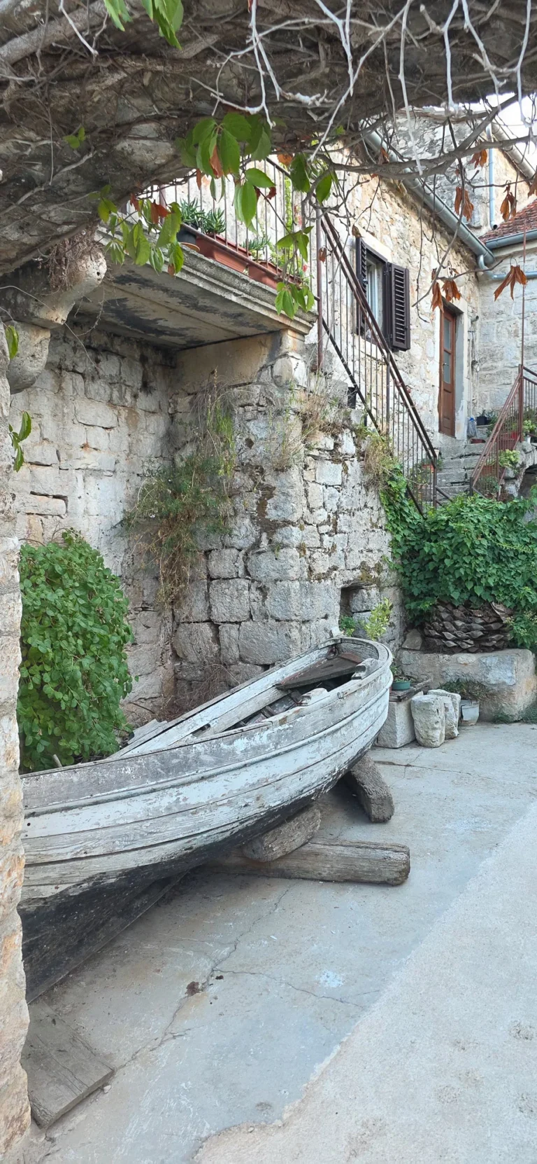 An old wooden boat rests beside a weathered stone house beneath overhanging tree branches.