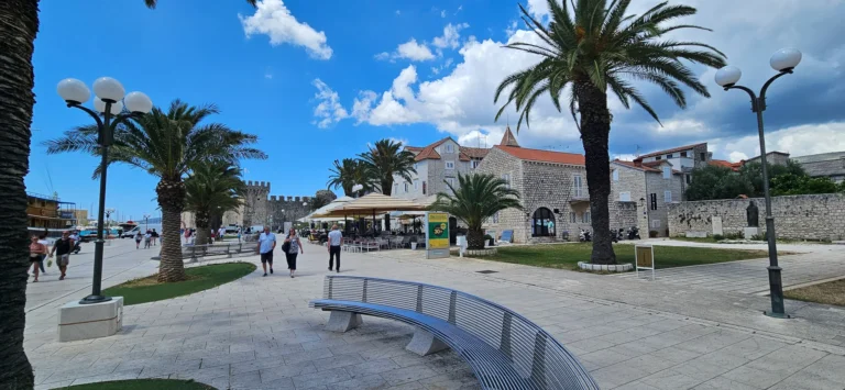 Paved coastal square with palm trees, stone buildings, a curved bench and people walking beneath a blue sky