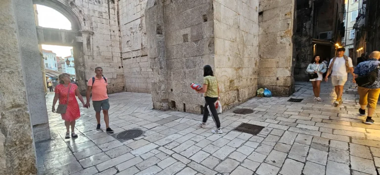 Pedestrians cross a stone-paved passage between old limestone buildings in Croatia.