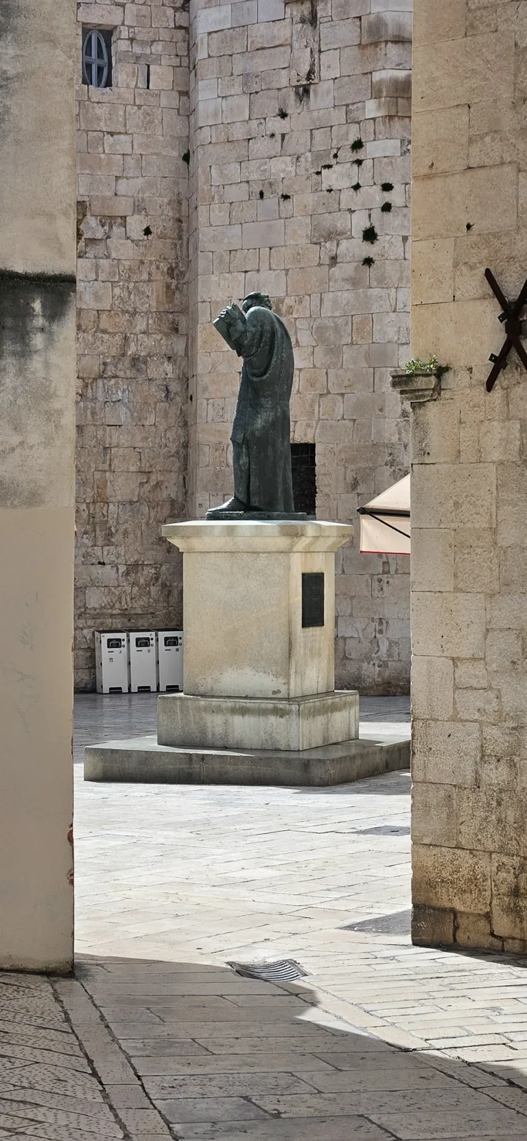 Bronze statue of a robed monk on a stone pedestal in a paved square framed by old stone walls