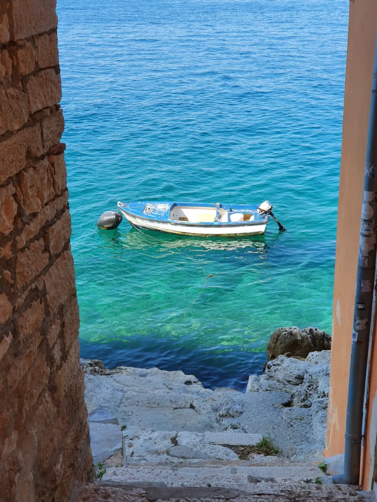 A small blue-and-white boat floats offshore beyond stone steps leading down to clear water.