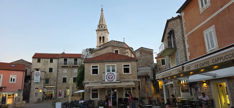 Stone buildings and café terraces surround a town square in Croatia, with a church bell tower rising behind them.