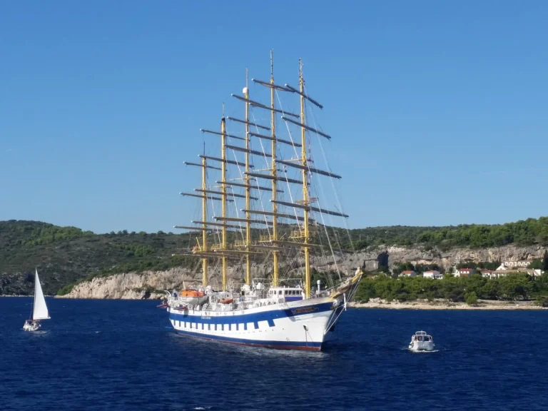 A tall sailing ship and smaller boats on blue water below a rocky, green Croatian coastline.