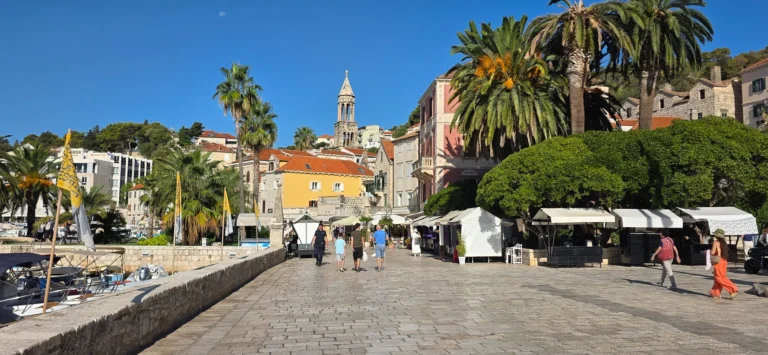 A stone-paved waterfront square with market stalls, palm trees, a bell tower and boats in the harbour.