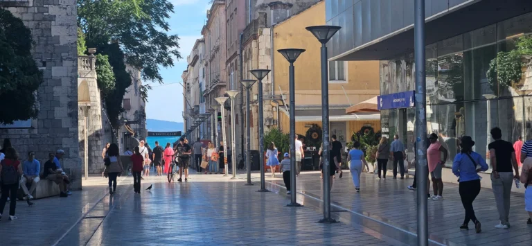 People walk along a paved pedestrian street lined with shops, older buildings and modern street lamps.