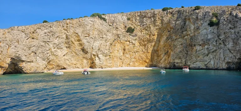 Small boats float below sunlit rocky cliffs beside a clear coastal cove.