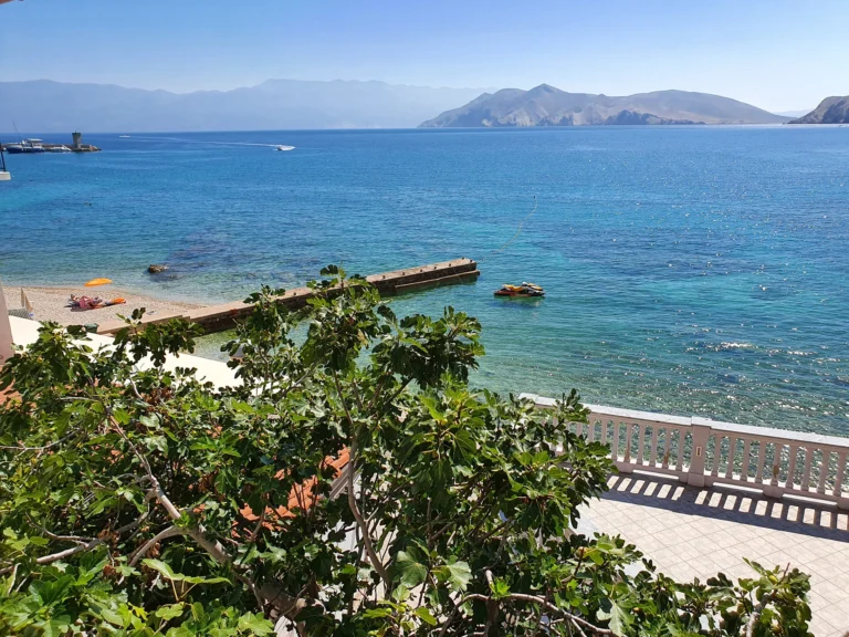 A small pebble beach and clear blue sea, with mountainous coastline across the water.