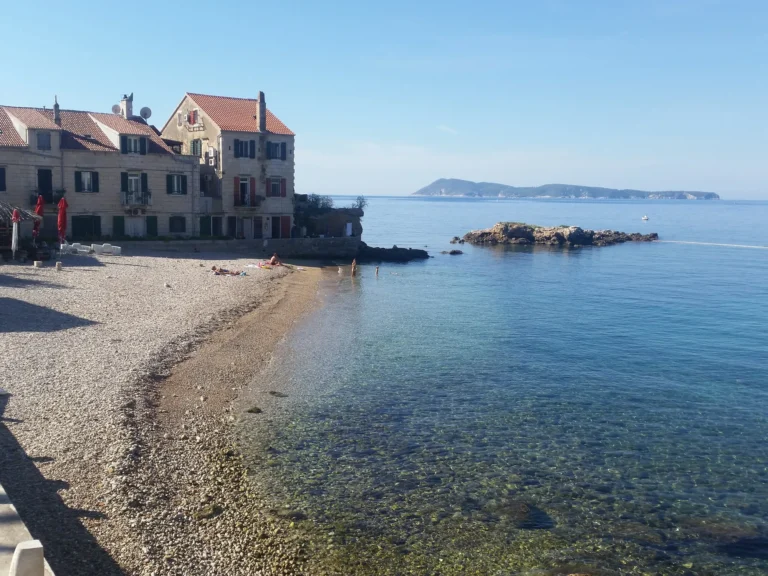 A pebbly beach and stone waterfront houses beside clear, calm water on the Croatian coast.