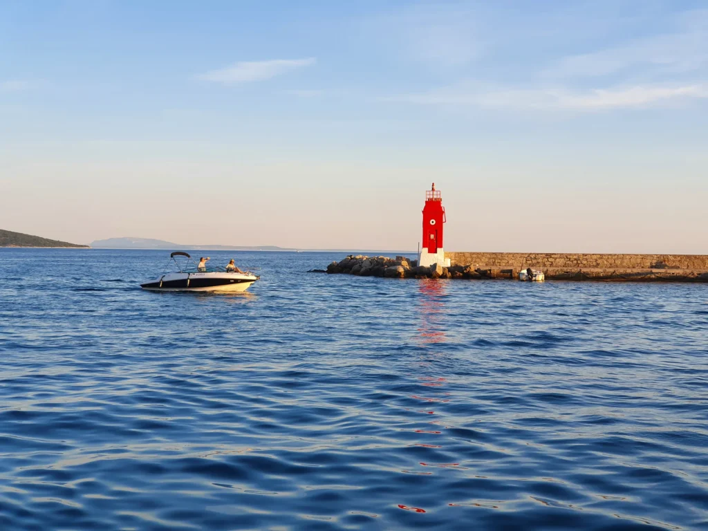 A red beacon marks a stone harbour entrance as a small motorboat crosses calm blue water.
