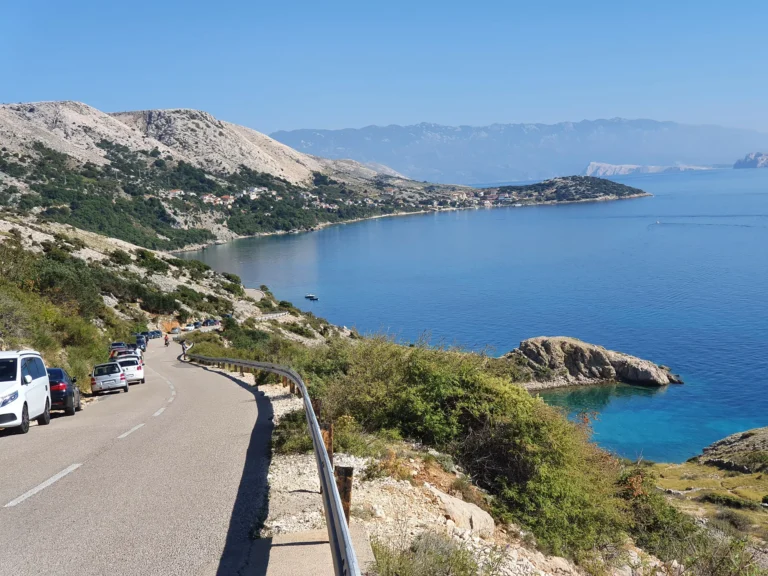 A winding coastal road overlooks a blue bay, rocky hills and scattered houses.