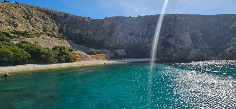 Turquoise water laps a narrow pebble shore beneath rugged coastal cliffs.