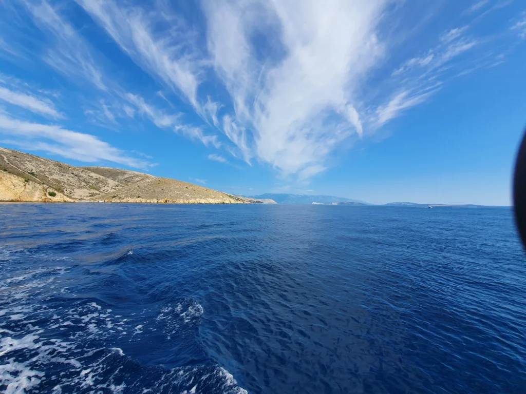 Deep blue sea and a pale rocky shoreline beneath wispy clouds, viewed from a boat.