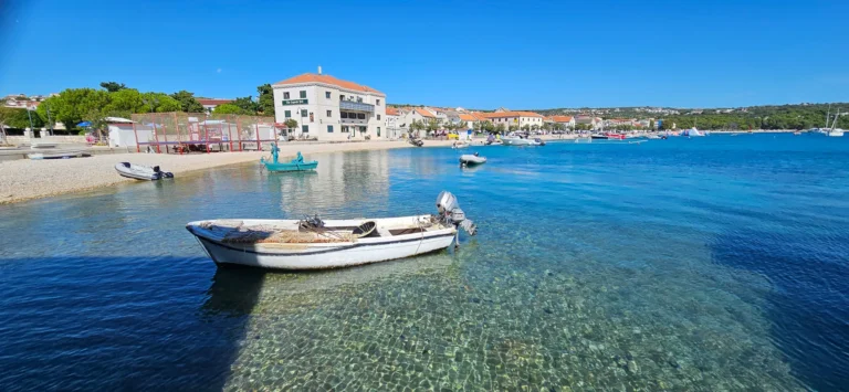 A small motorboat floats in clear water beside a Croatian town beach and waterfront buildings.